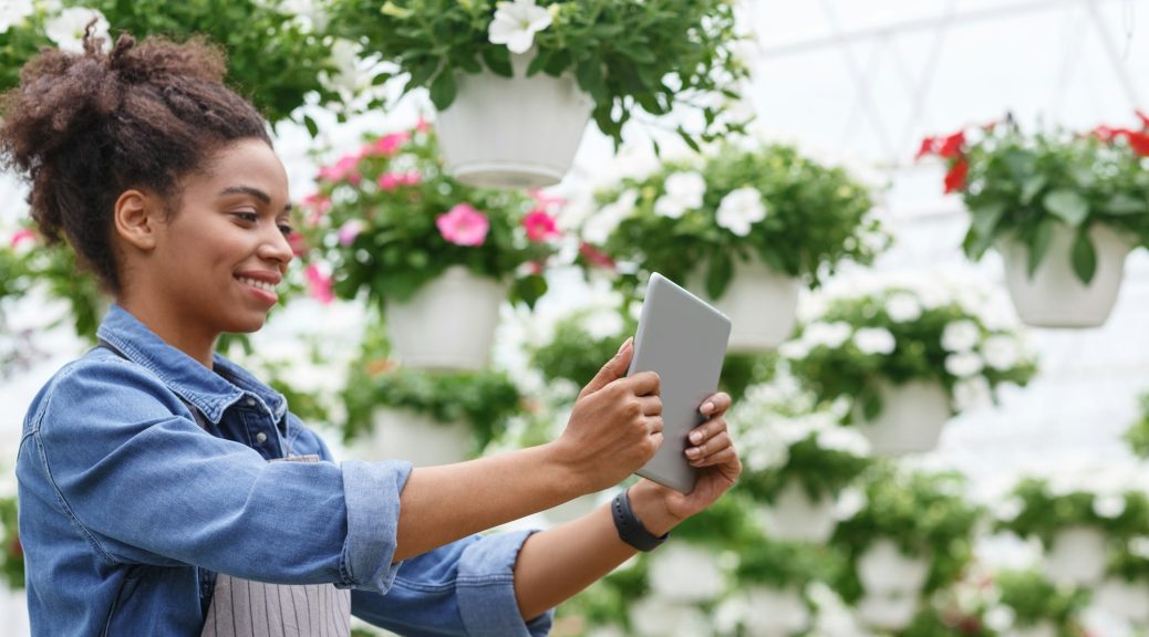 Working with digital technology in greenhouses with flowers. African american and farm worker girl